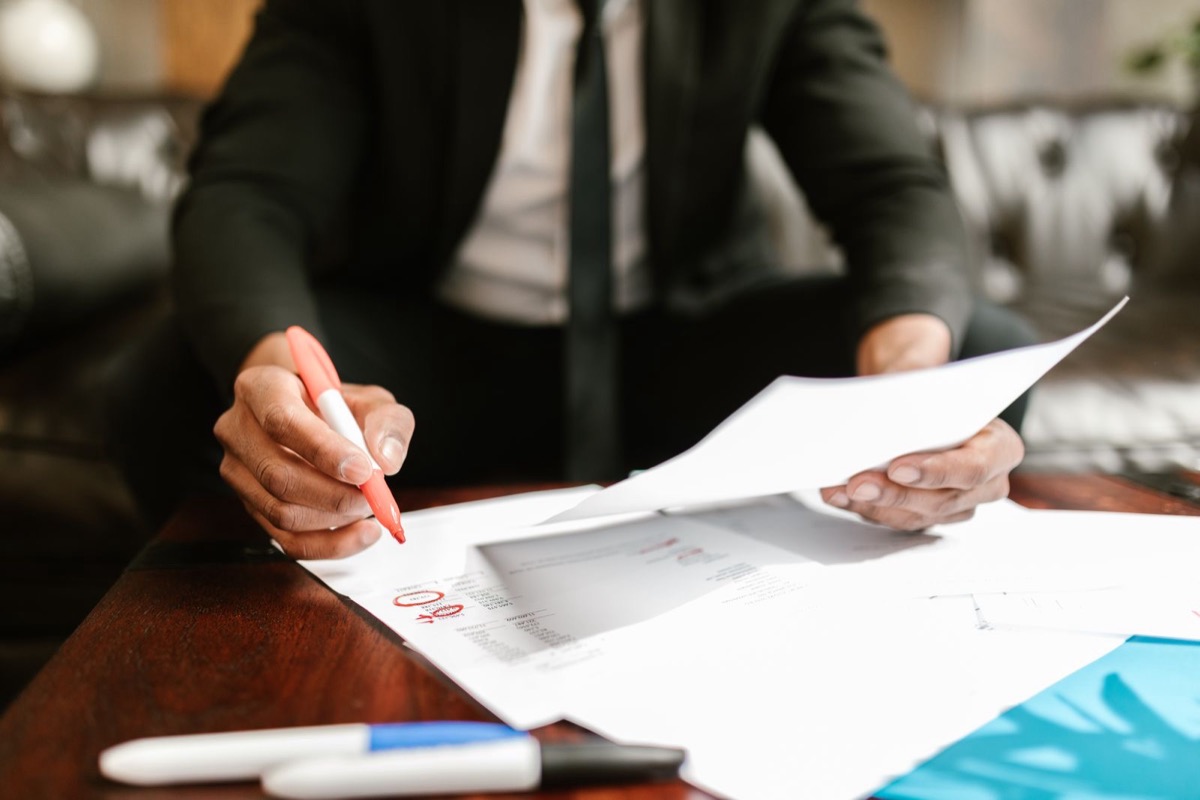 A hand holding a red pen over printed pages on a dark wooden desk, a blue folder and two markers at the edge