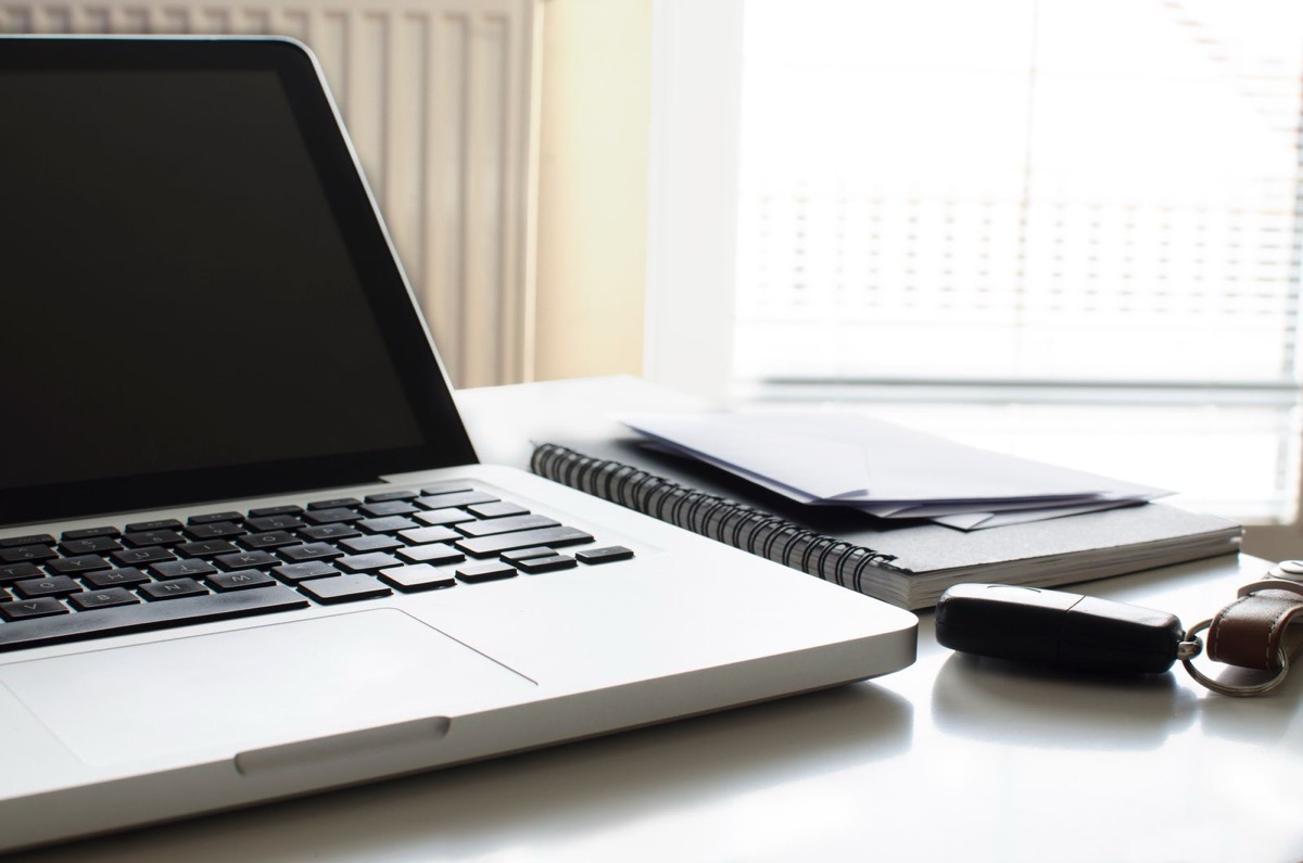 A silver laptop with a dark screen beside a spiral notebook topped with envelopes and a car key, on a white desk by a window