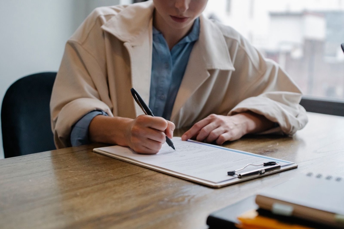 A person in a beige coat, face out of frame, signing a form on a clipboard at a wooden desk