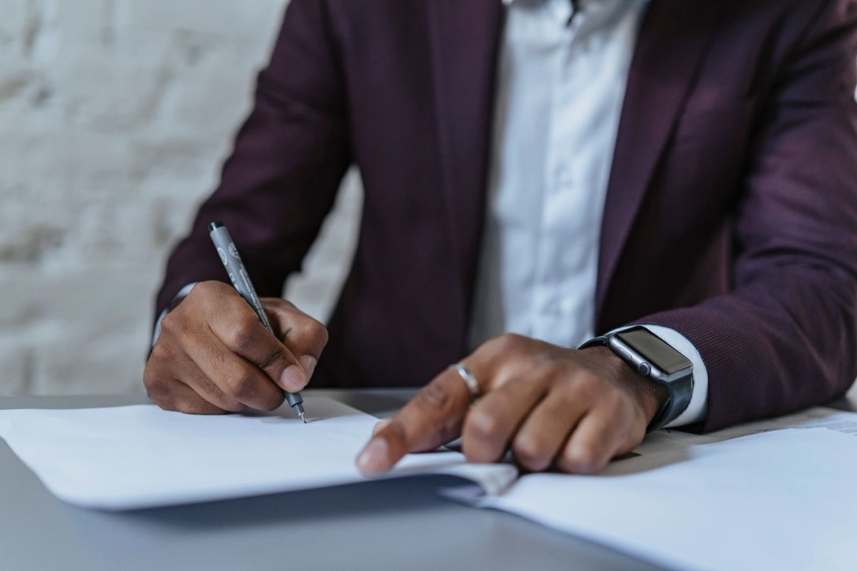 A man in a dark suit, seen from the chest down, signing a sheet of paper at a desk with a black pen