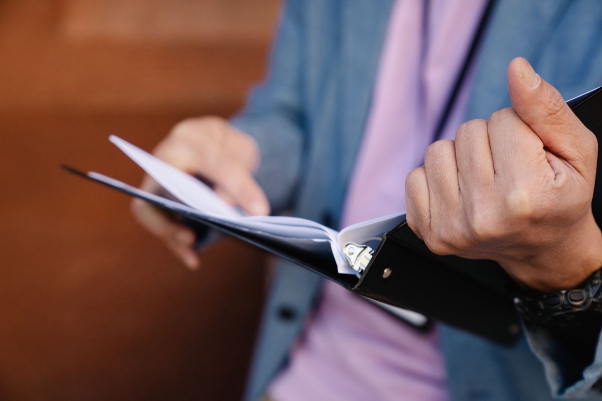 Hands holding open a black ring binder, a person in a blue jacket and lilac shirt seen from the chest down