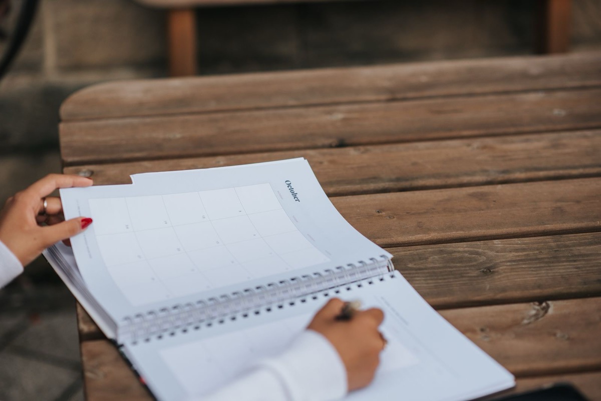 A hand writing in an open spiral planner with a blank monthly grid on a wooden table
