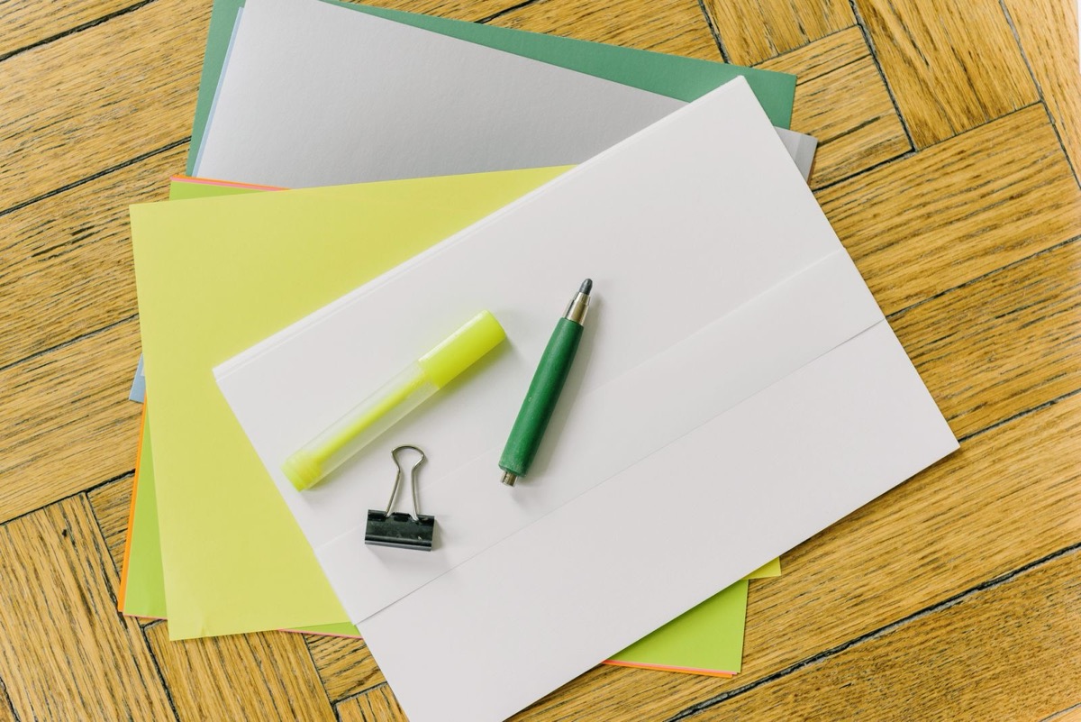 A stack of white, yellow and green sheets on a wooden parquet surface with a highlighter, a pen and a binder clip, seen from above