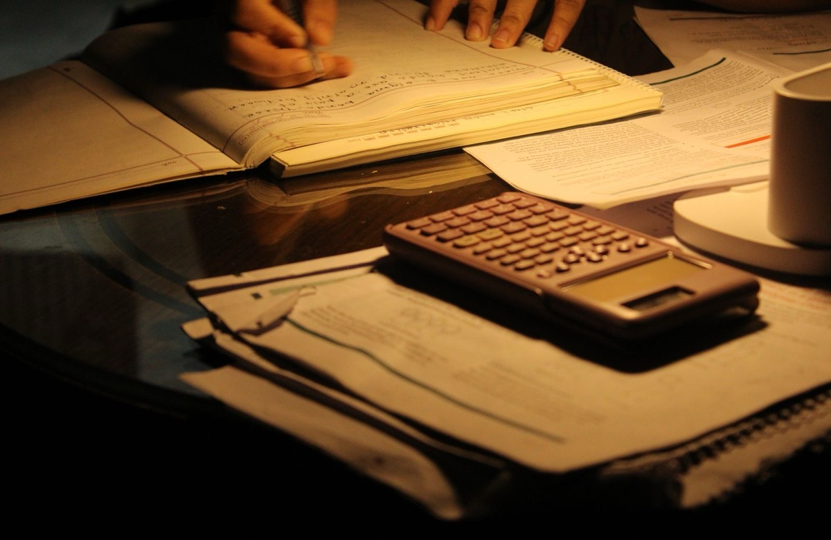 A calculator on stacked papers in warm lamp light, with a person's hands writing in a ledger behind it