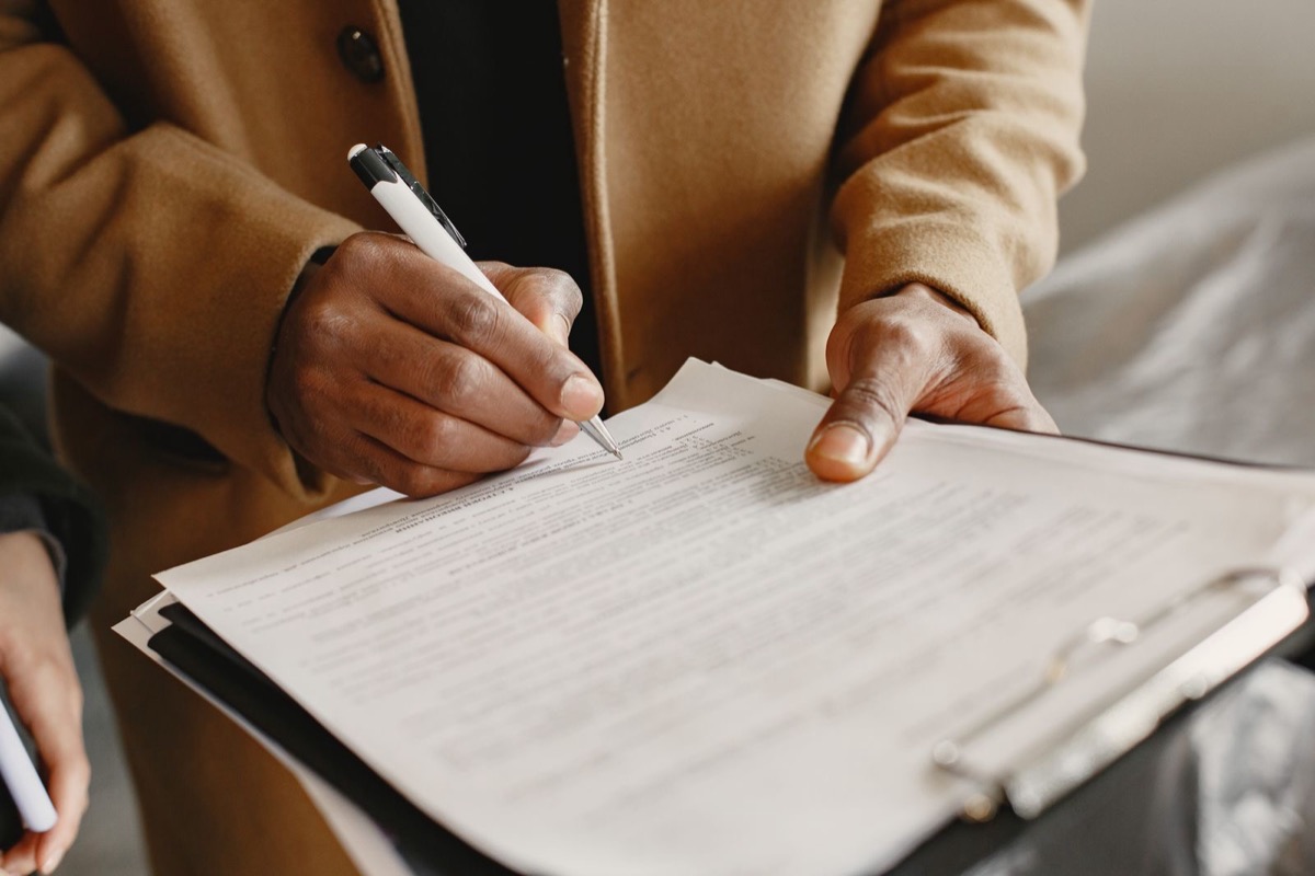 A person in a tan coat signing printed pages on a clipboard with a white pen