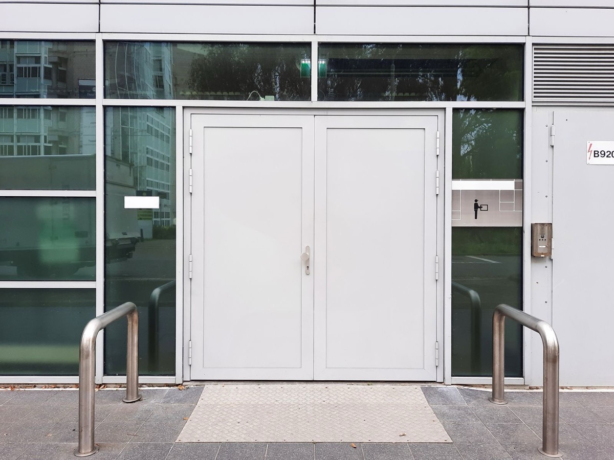 White double doors in a glass-fronted building, with steel bollards on the pavement in front