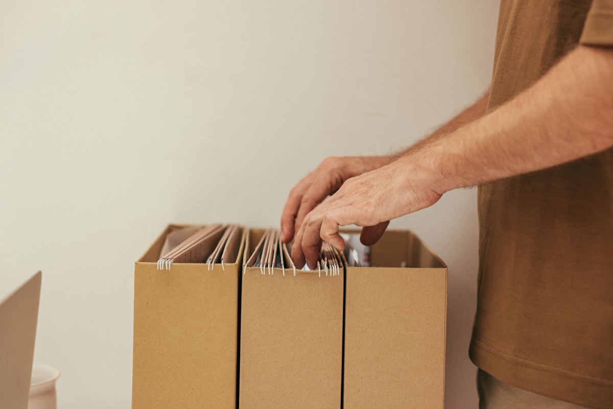 A hand filing hanging folders into a row of cardboard boxes against a pale wall