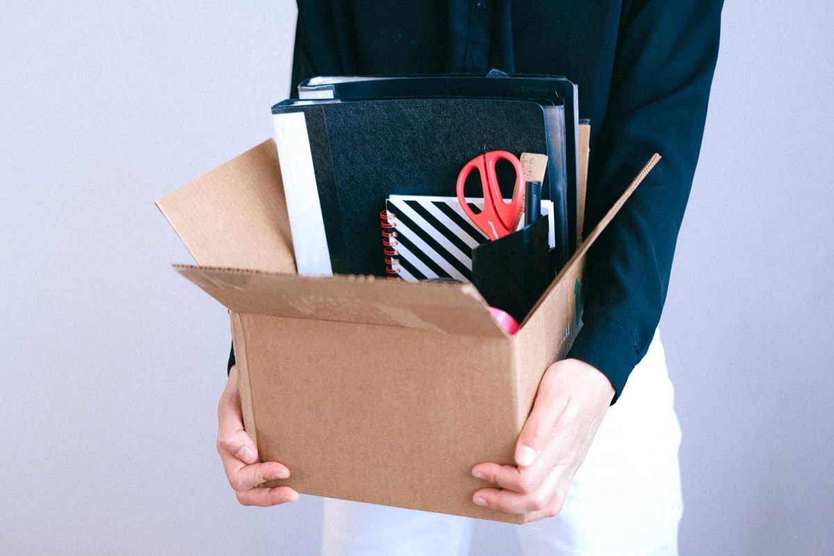 A person holding a cardboard box of folders, a notebook and red scissors, seen from the chest down