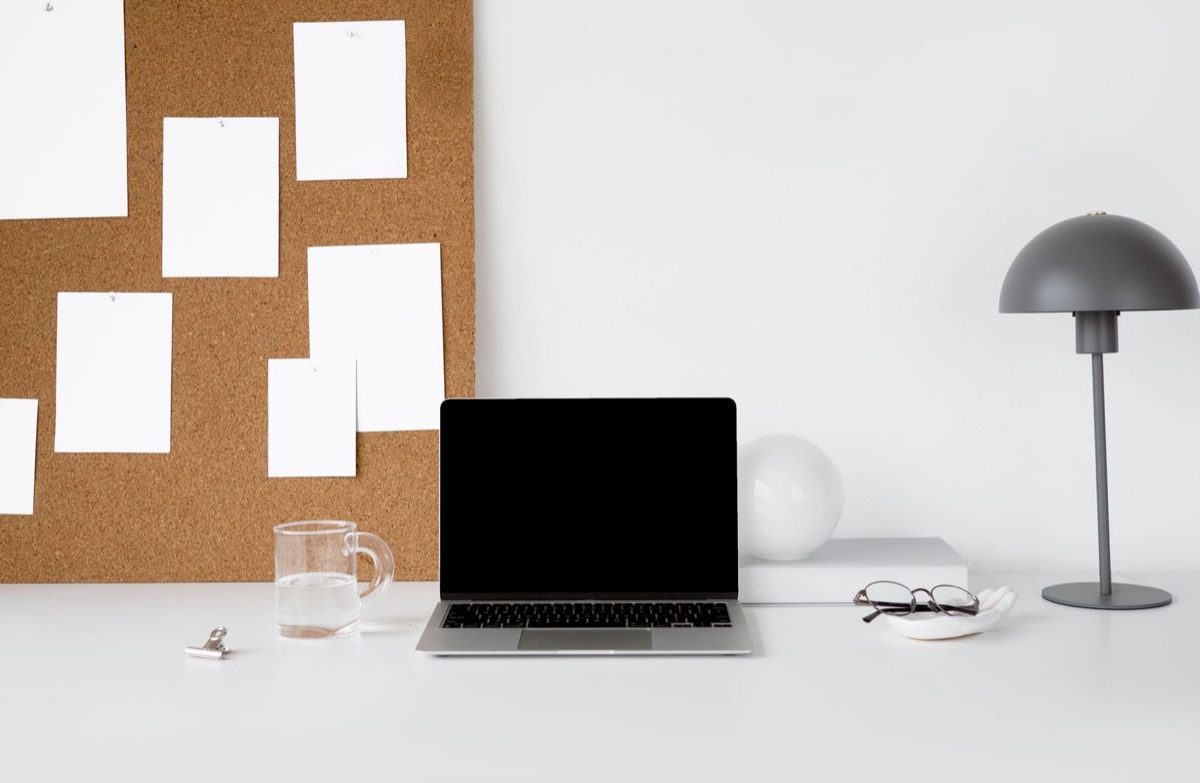 A laptop with a black screen on a white desk in front of a cork board with blank white cards, beside a lamp, a glass of water and a pair of glasses
