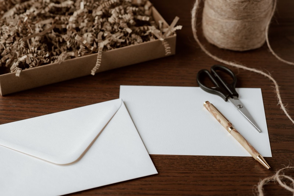 White envelopes, a pen and scissors on a dark wooden desk beside a cardboard box of paper shreds and twine