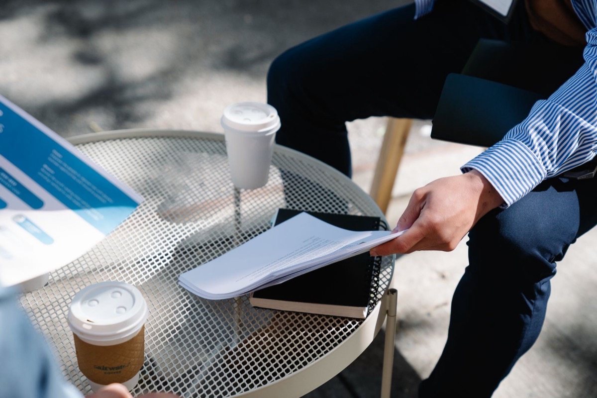 A man's hands holding loose printed pages above a round table with takeaway coffee cups, seen from the waist down