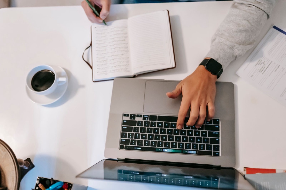 Hands on a laptop keyboard beside an open notebook, a cup of coffee and a printed sheet on a white desk, seen from above