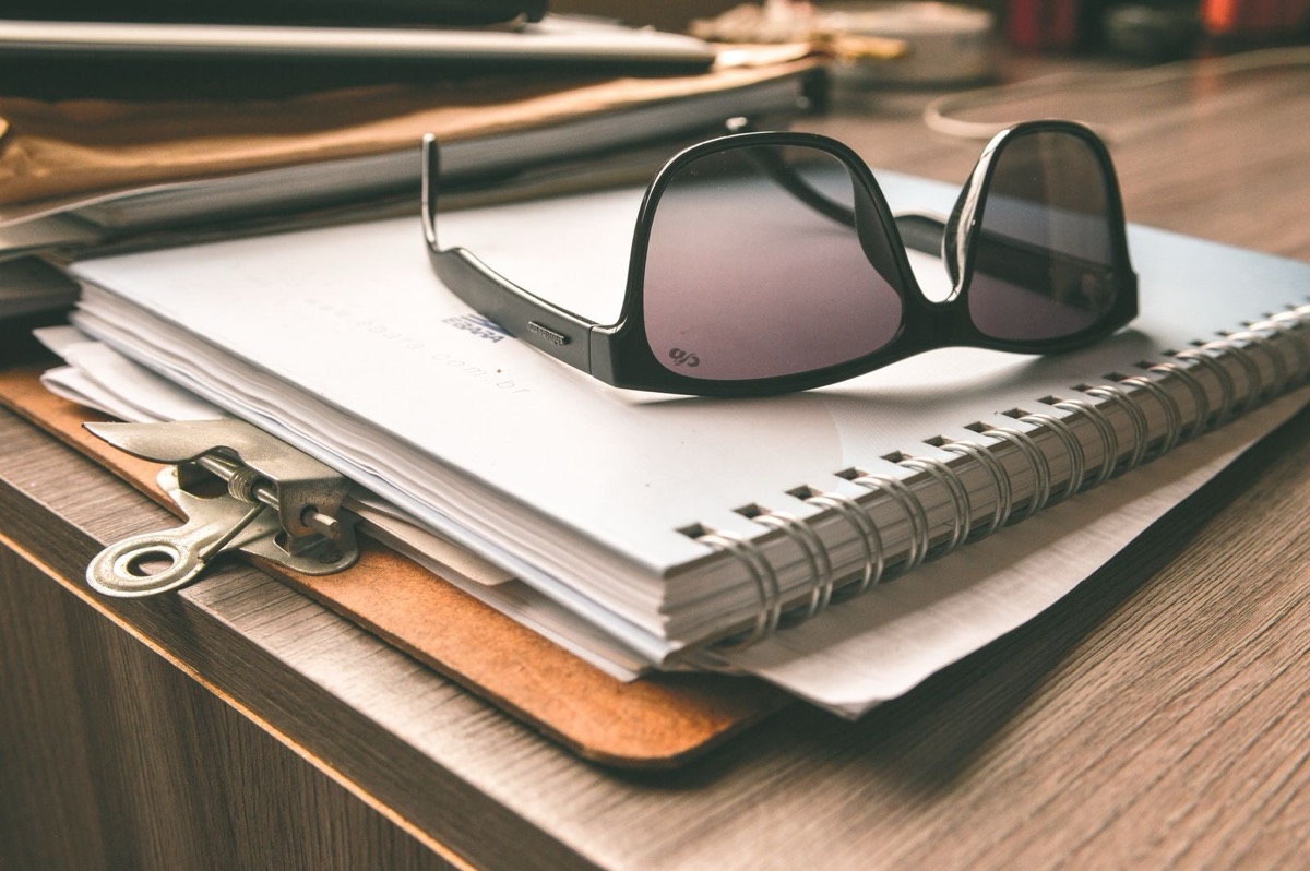Sunglasses on a spiral notebook and papers on a clipboard on a wooden desk