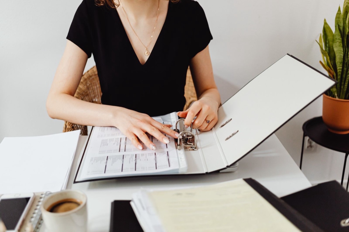 A person's hands on an open ring binder of printed tables, with a cup of coffee and other binders on a white desk