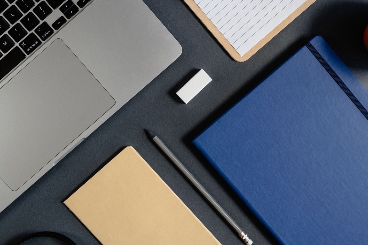 A laptop corner, a blue notebook, a kraft notebook and a pencil on a dark desk, seen from above