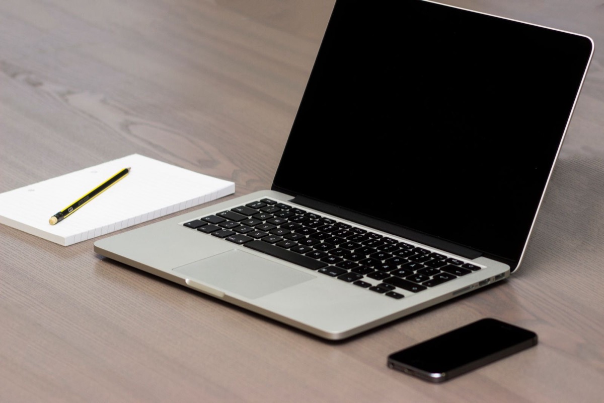 A laptop with a blank screen, a notepad with a pencil and a phone on a pale wooden desk