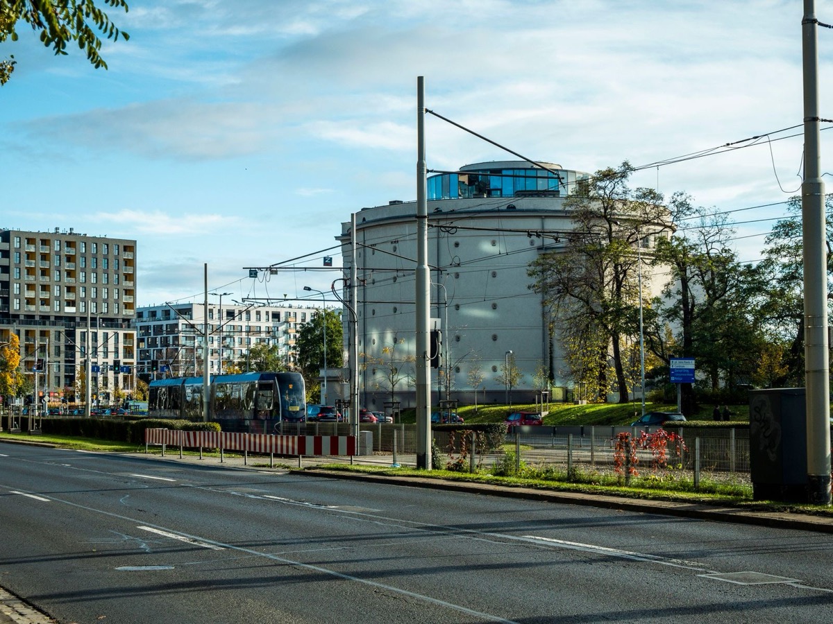 A city road with tram wires and a tram in the distance, apartment and office buildings and trees under a partly cloudy sky