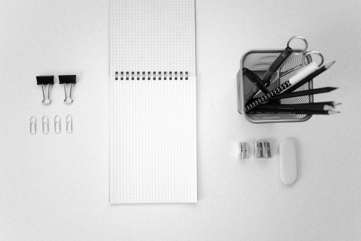 A spiral notepad, black binder clips, paper clips and a pen cup on a pale desk, photographed in black and white