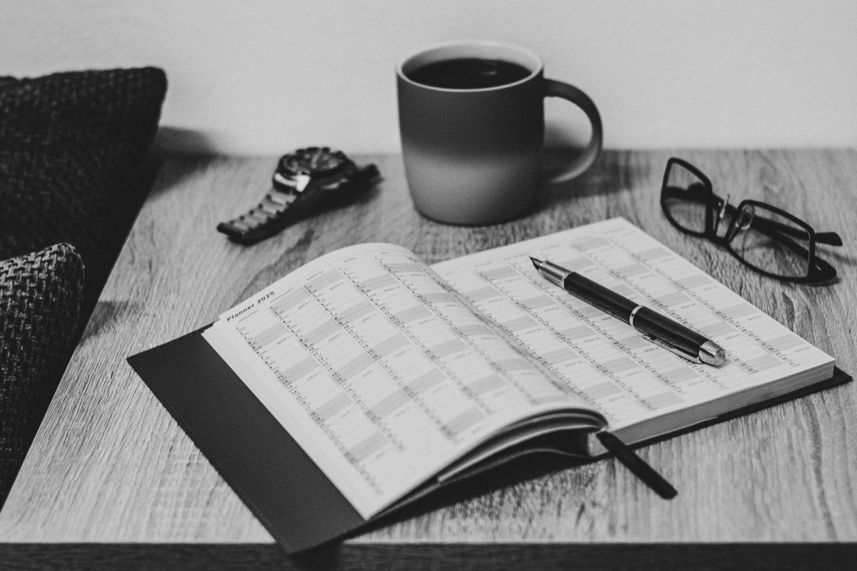 A black-and-white view of an open planner with a pen, glasses, a watch and a mug on a wooden desk