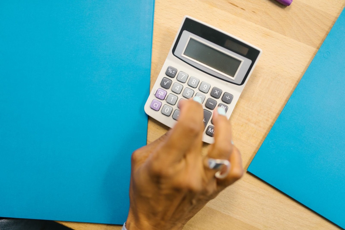 A hand pressing the keys of a calculator on a blue folder on a wooden desk