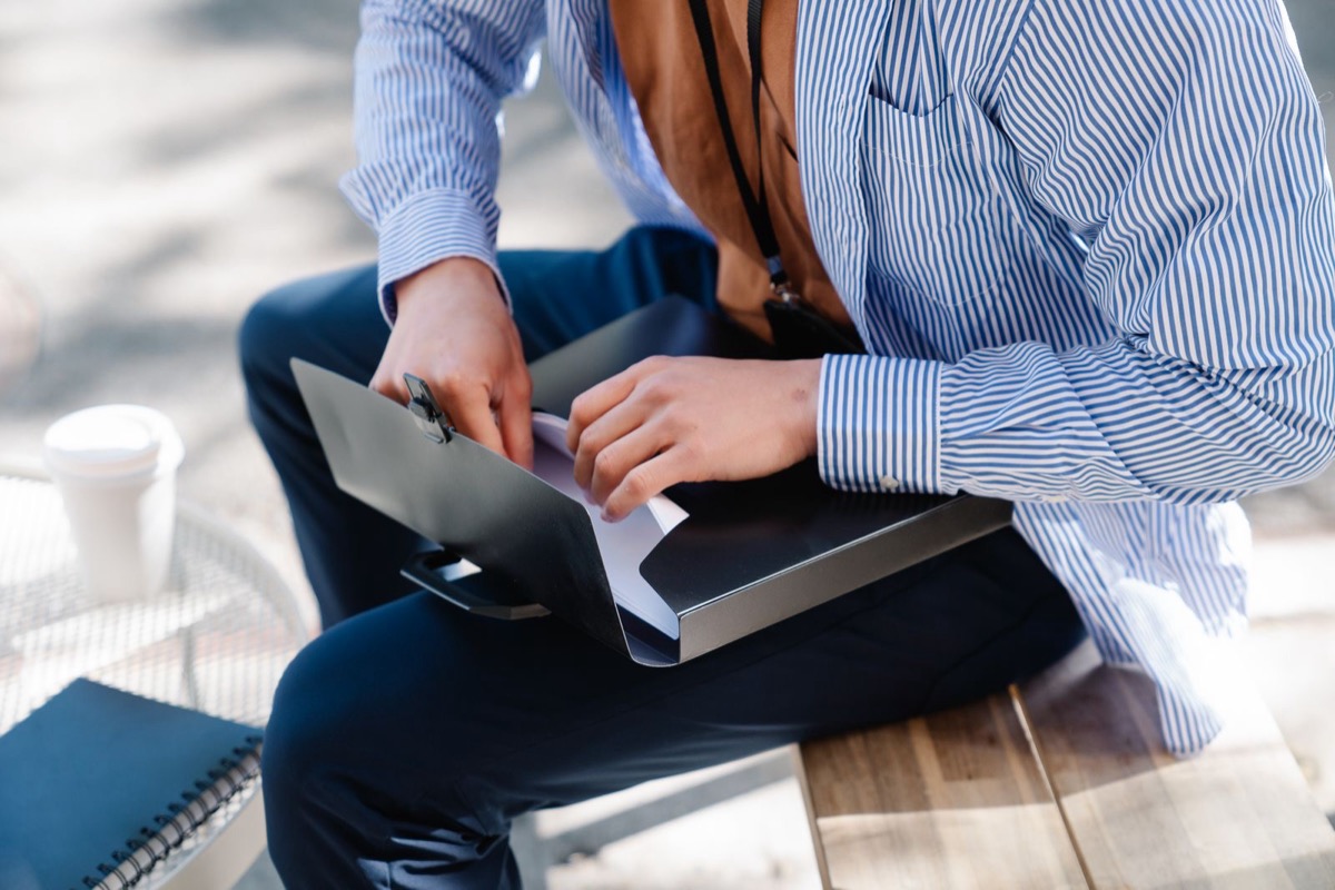 A person seen from the chest down, seated on a bench with an open black folder of papers on their lap