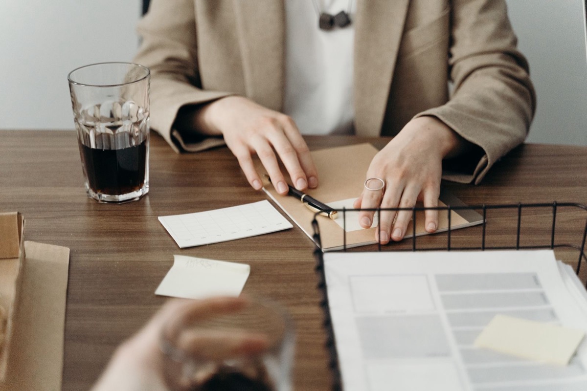 Hands resting on a kraft folder with a pen on it, beside a glass of dark drink, note cards and a wire tray of papers on a wooden desk