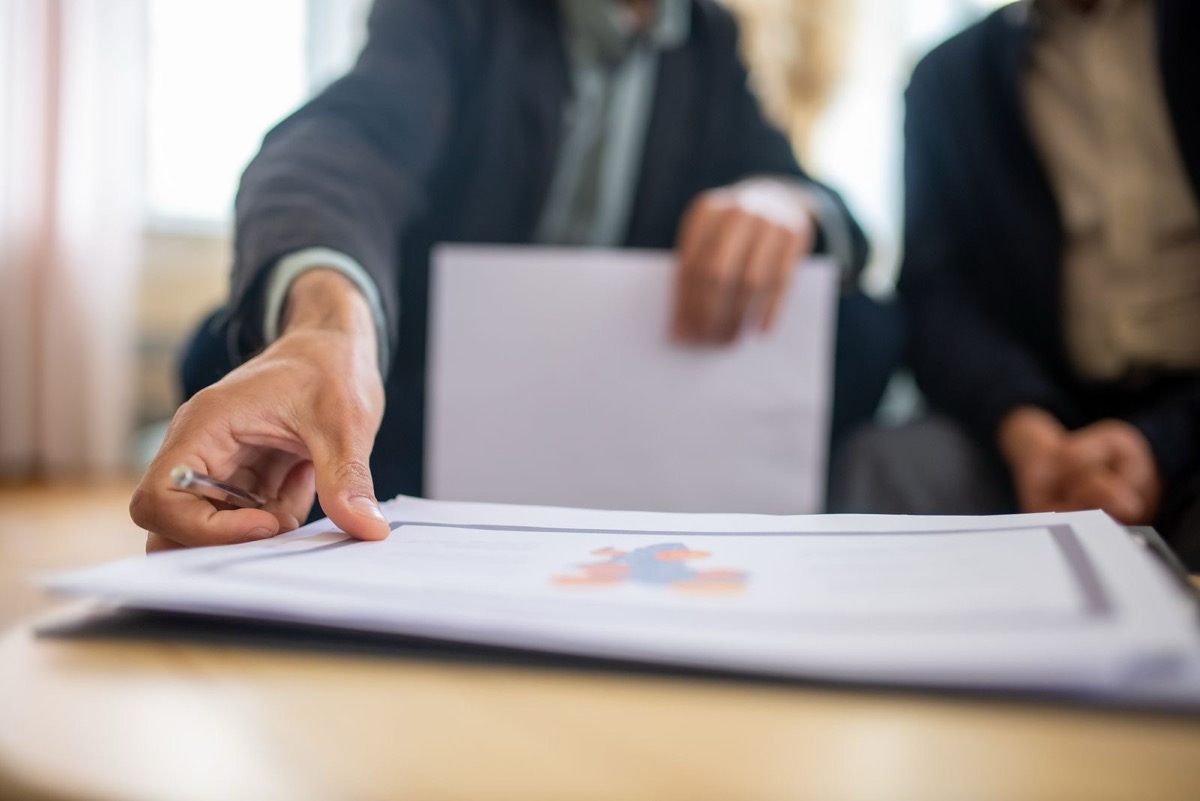 A hand with a pen pointing at a printed page with a small chart on a desk, two seated people in dark suits blurred behind