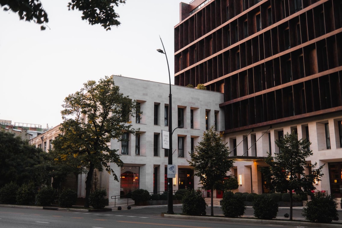 A quiet street between a pale stone building and a dark modern office block, with trees