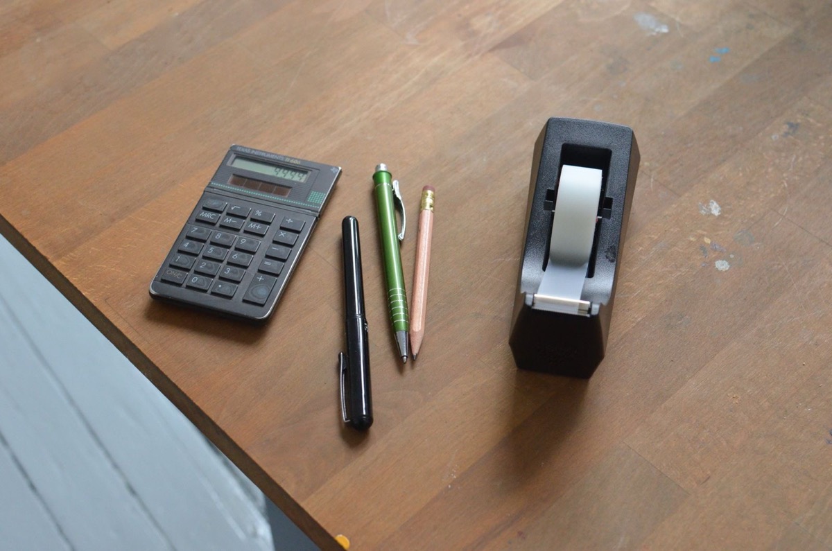 A pocket calculator, three pens and a tape dispenser on a wooden desk