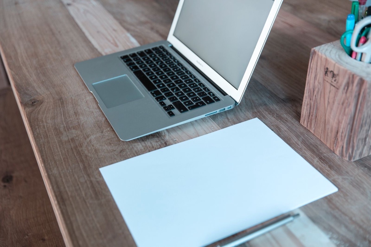 An open silver laptop with a pale screen on a wooden desk beside a pale-blue sheet of paper and a pen