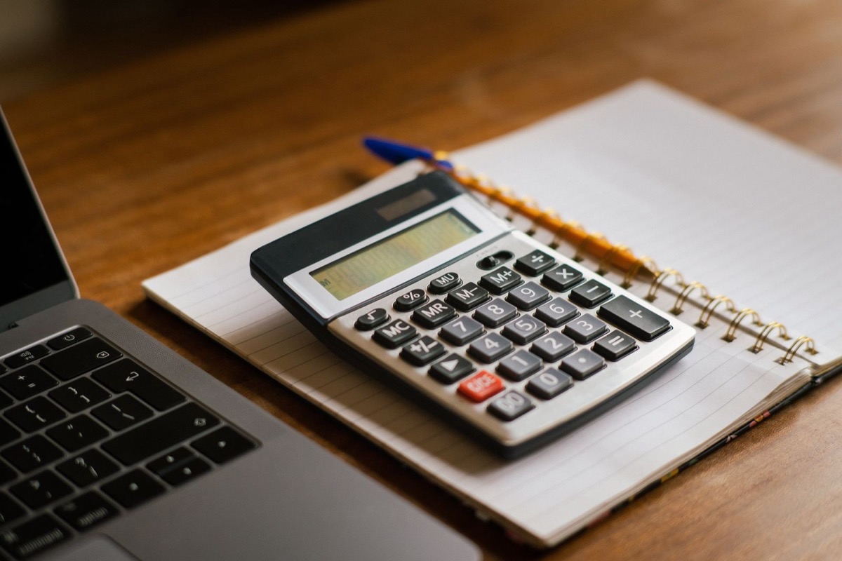 A calculator on an open spiral notebook with a pencil and a blue pen, the edge of a laptop at the left