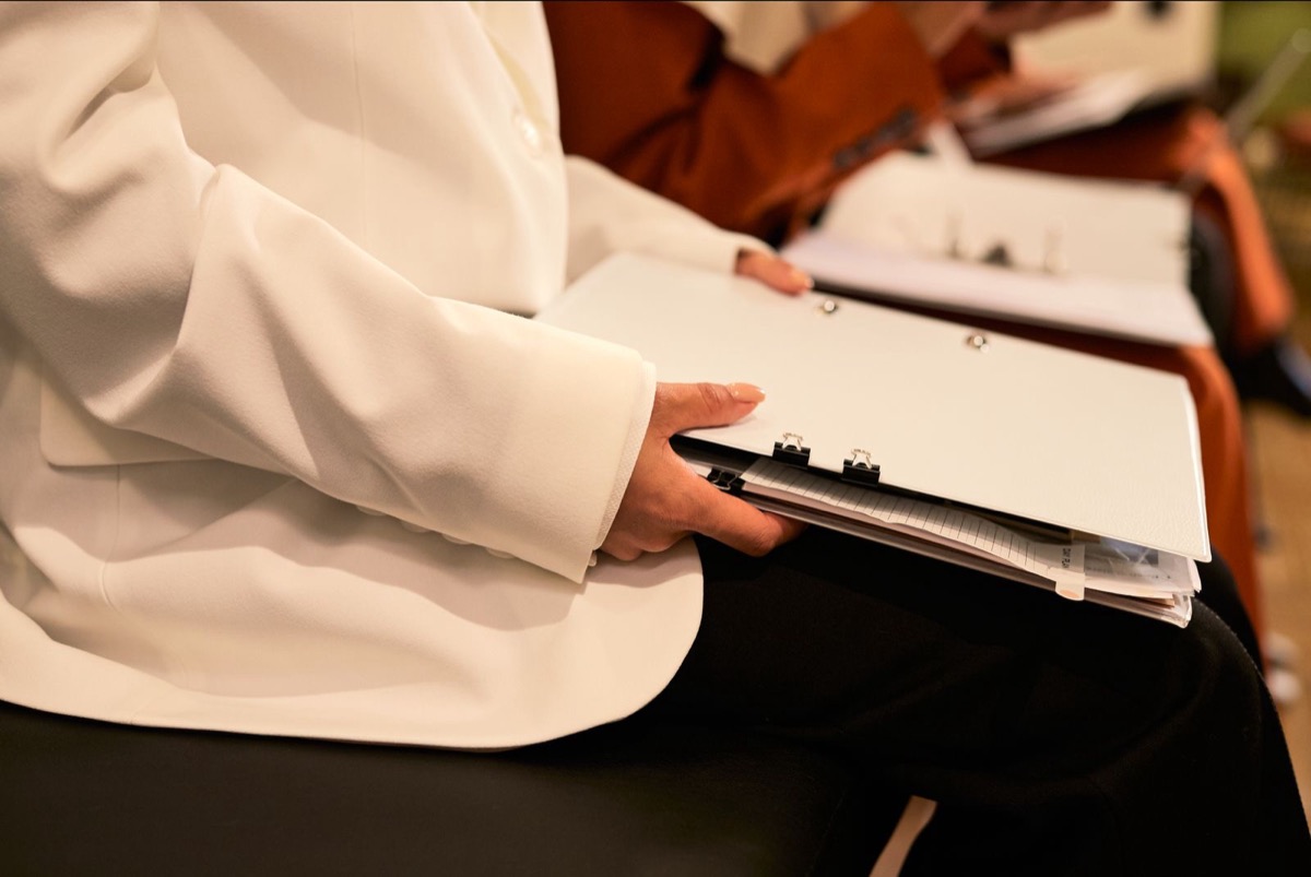 A woman in a white jacket seated with a binder and loose papers on her lap, other people seated beside her