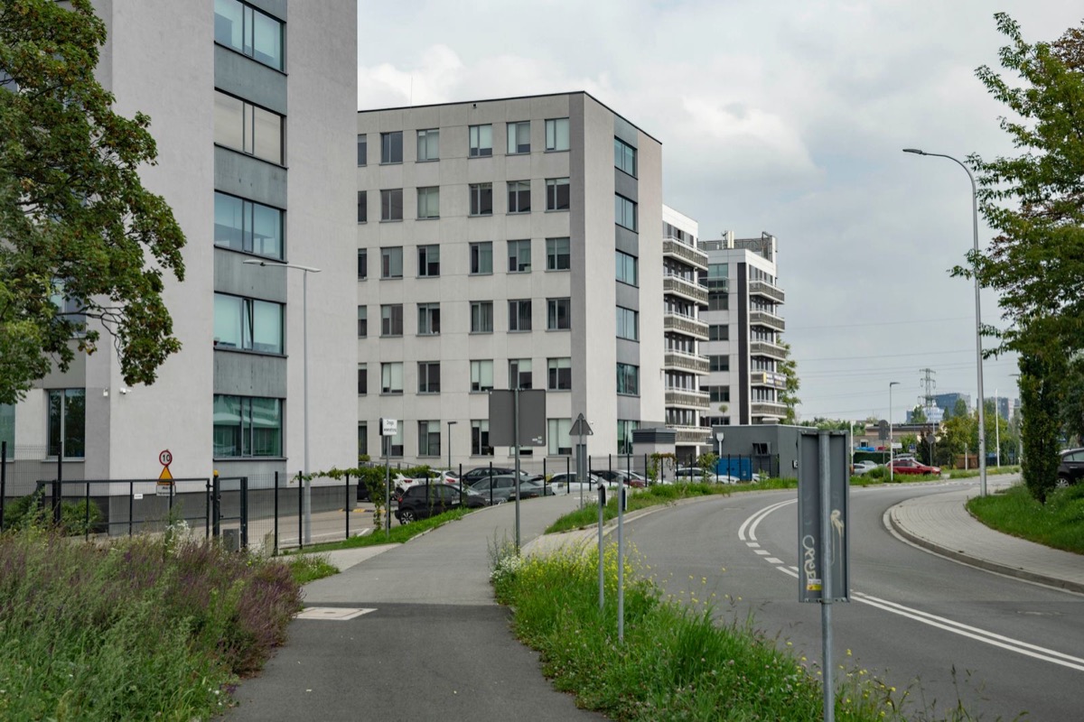Grey office and apartment buildings beside a curving road with a footpath, grass and trees under a cloudy sky