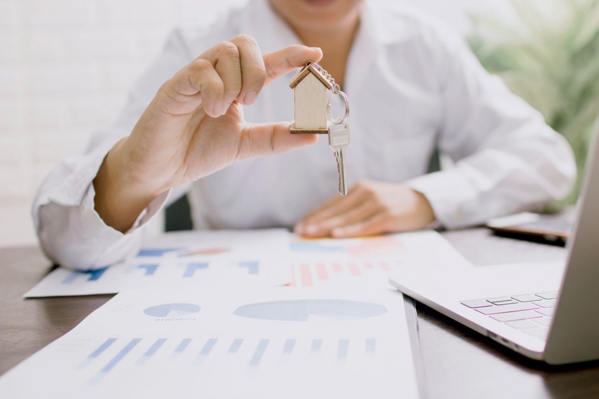 A hand holding a set of keys on a house-shaped tag above printed charts on a desk, with a laptop at the edge