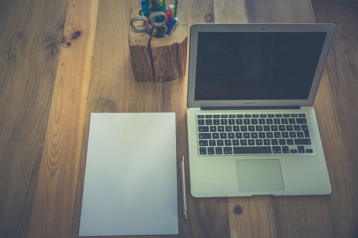 An open laptop with a dark screen beside a blank sheet of paper and a pen on a wooden desk