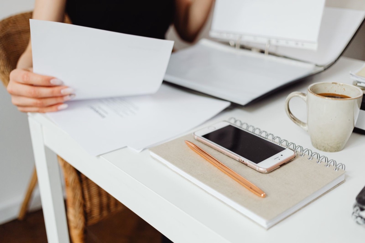 A person's hands holding a sheet of paper over a white desk with an open ring binder, a notebook, a phone, a pen and a mug of coffee