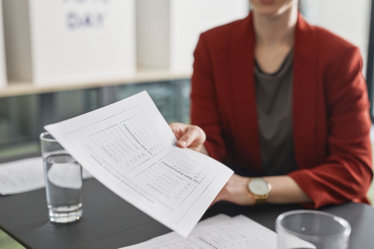A woman in a red jacket, face out of frame, holding a printed page with a table across a dark desk, two glasses of water in front of her
