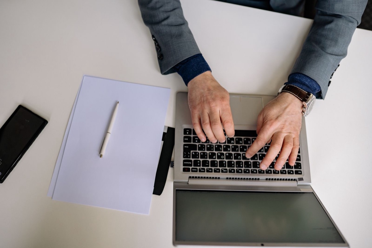 Hands typing on a laptop beside a blank notepad, a pen and a phone, seen from above on a white desk