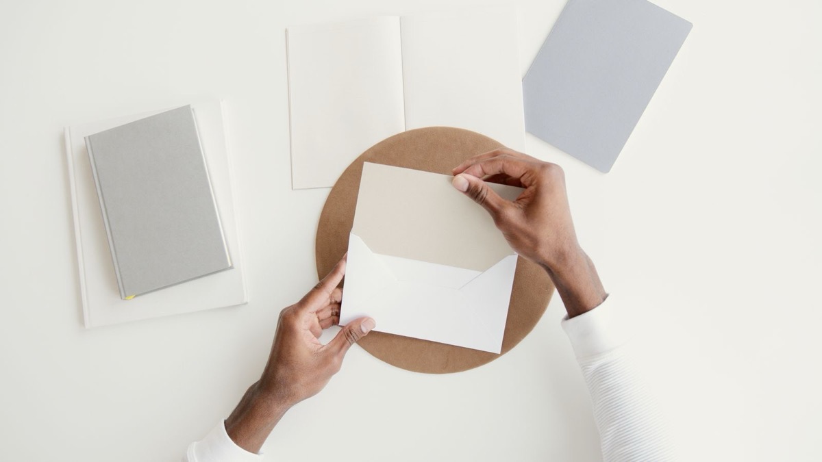 Two hands opening a white envelope on a clean desk beside a closed grey notebook and blank cards
