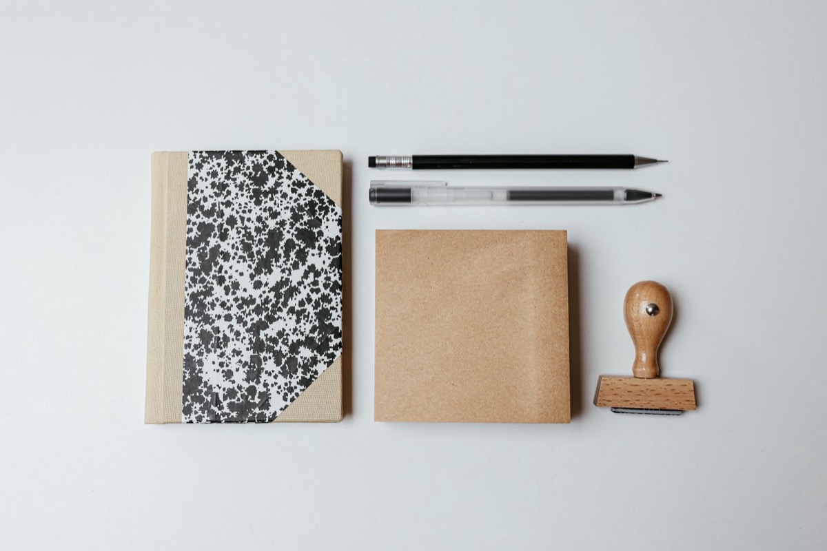 A wooden-handled rubber stamp beside a notebook, a kraft envelope and two pens on a pale desk
