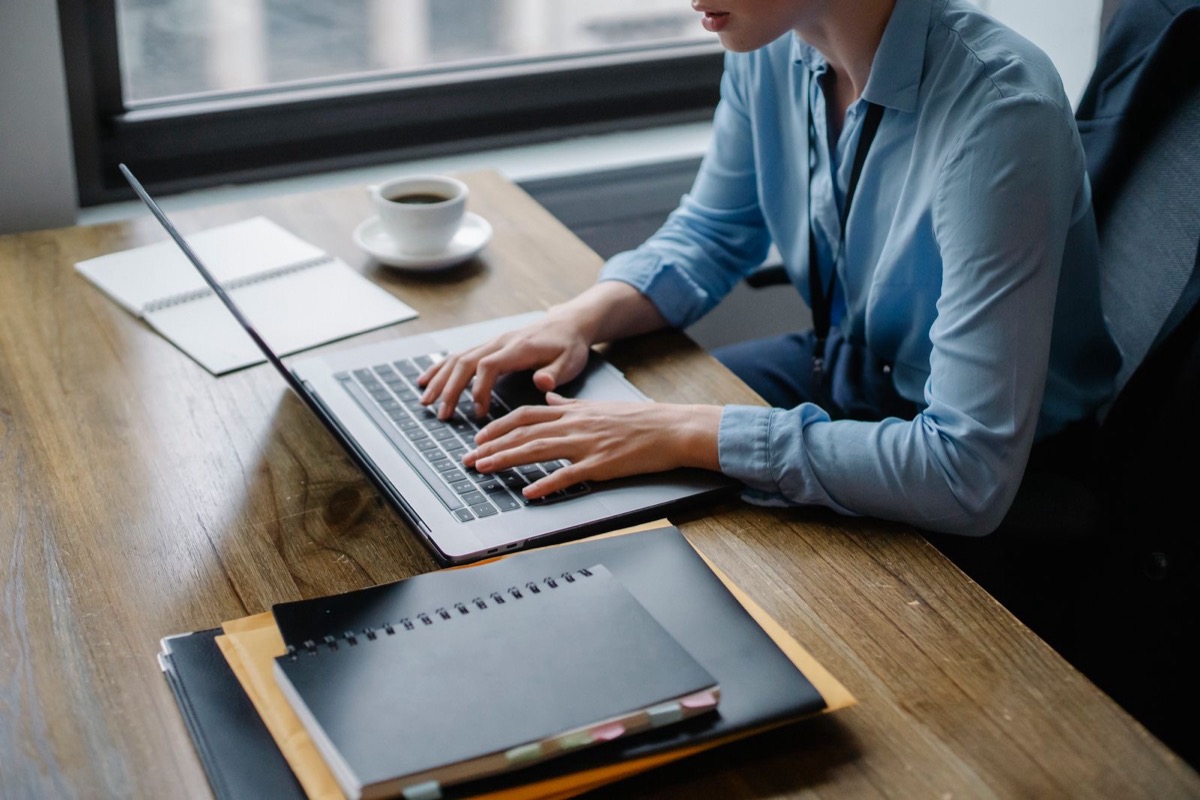 A person in a pale blue shirt, face out of frame, typing on a laptop at a wooden desk by a window, with a notebook, folders and a cup of coffee