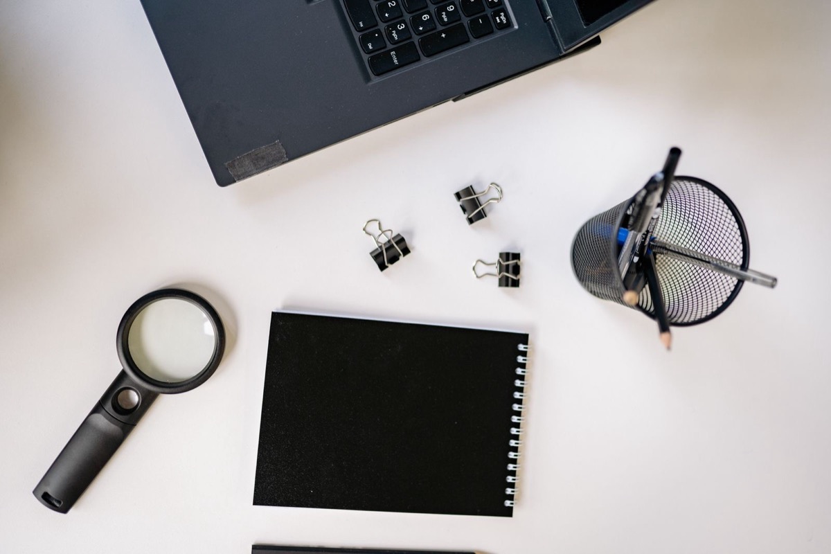 A black spiral notepad, a magnifying glass, binder clips and a pen cup on a white desk beside the edge of a laptop