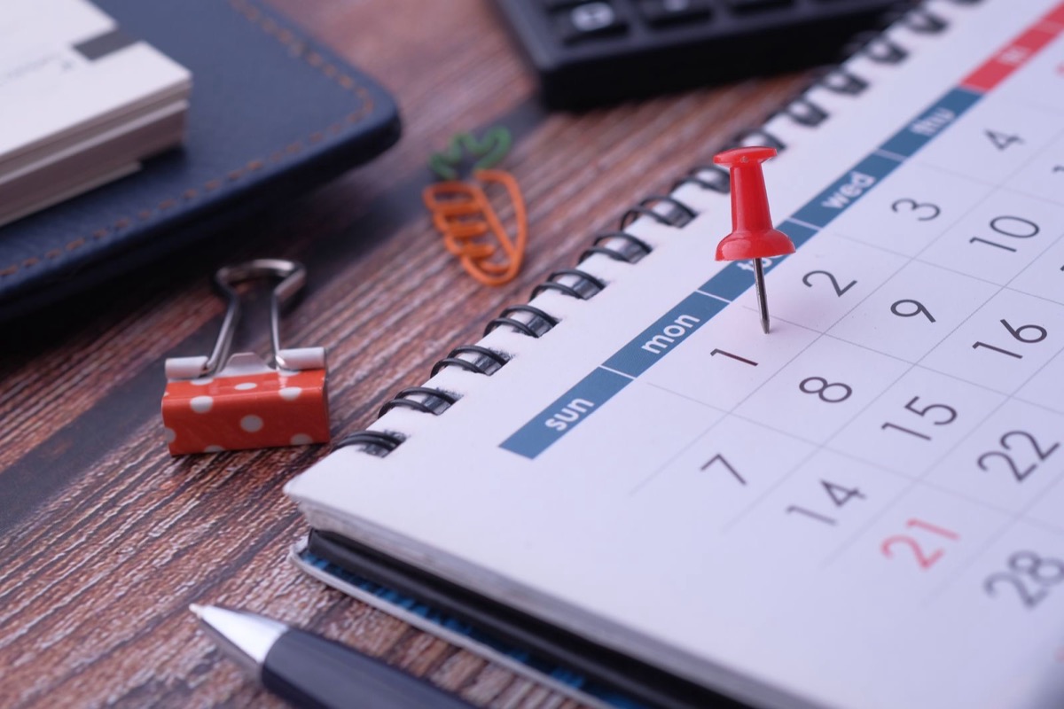 A desk calendar with a red pin on one date, a binder clip and a pen on a wooden desk