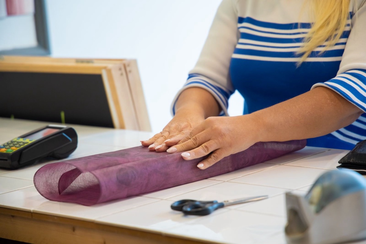 A shop assistant's hands wrapping purple tissue on a counter beside a card terminal and scissors
