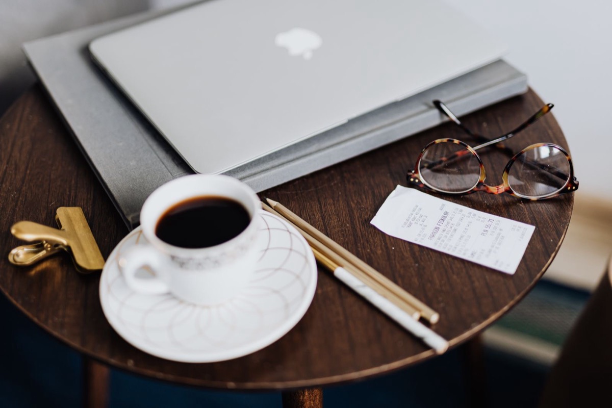 A closed laptop on a grey folder beside a cup of coffee, glasses, pencils and a small receipt on a round wooden table
