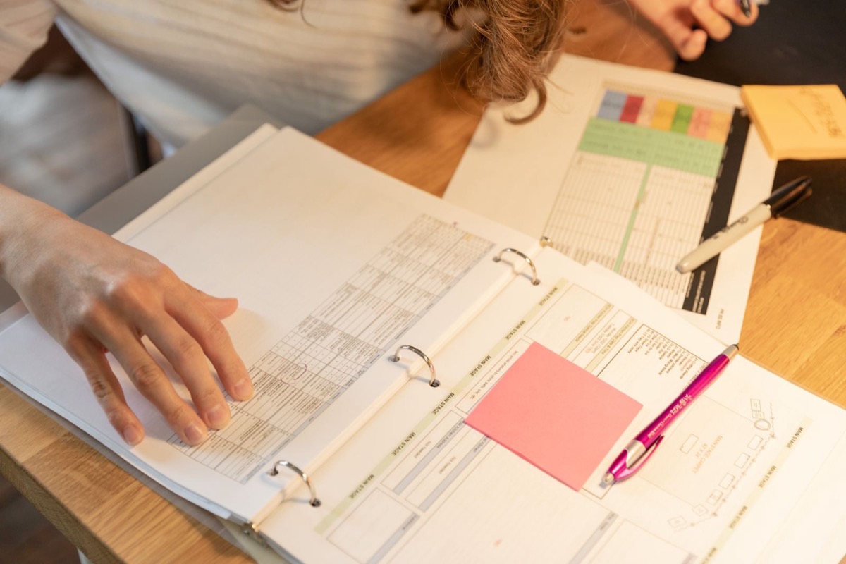 A hand resting on an open ring binder of printed tables beside a pen and a sticky note