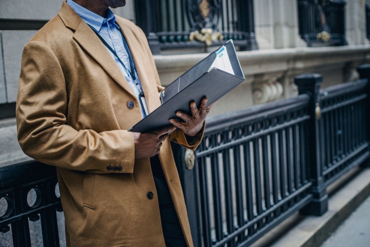 A man in a camel coat, face out of frame, opening a dark folder beside an iron railing